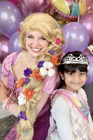 A light-skinned woman wearing a pink dress, with long braided blond hair woven with flowers. Next to her is a dark-haired girl with a medium skin tone wearing a dress and a silver tiara. Background is pink and purple balloons. Credit: Parties with Character