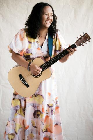 Photograph of a woman with dark hair and medium brown skin, wearing a floral dress and holding a guitar. She is laughing or smiling at something out of the shot.