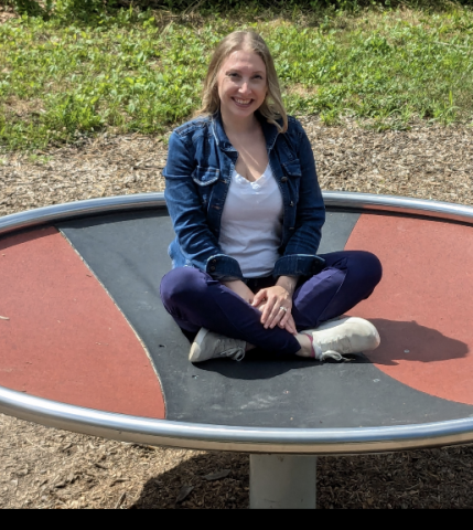 A light-skinned, blond woman wearing a denim jacket, white t-shirt, jeans, and white sneakers sits on a trampoline. The trampoline is on a grassy lawn. The woman is sitting cross-legged with her hands in her lap and smiling.