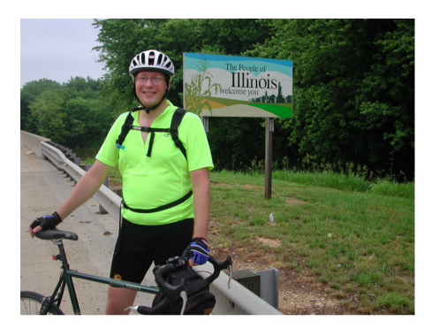 Jeff Spaetzel is dressed in cycling gear with his bike and stands by a Welcome To Illinois sign
