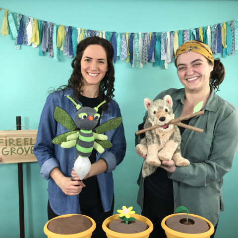 Two women in cardigans holding puppets, one a firefly and one a wolf pup, in front of a festive blue background.